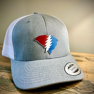May include: A gray and white trucker hat with a red, white, and blue state of South Carolina outline with a lightning bolt design. The hat has a Snapback closure.