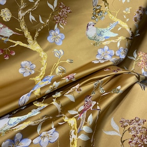 May include: Close-up of a golden-yellow fabric with an embroidered design. The design features birds perched on branches with white leaves, blue flowers, and red and brown accents. The fabric has a slight sheen.