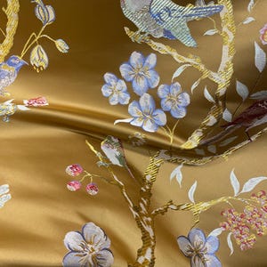 May include: Close-up of a golden fabric with an embroidered design featuring birds perched on branches with blue flowers. The fabric has a satin sheen and a detailed, textured pattern.