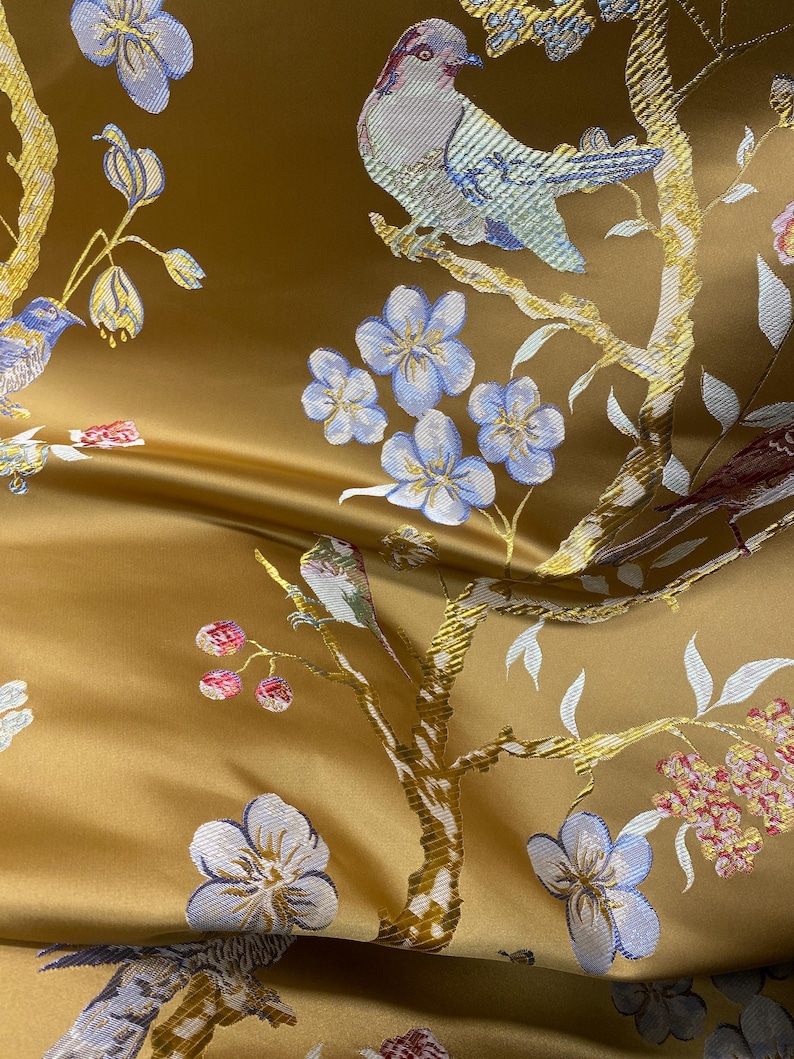 May include: Close-up of a golden fabric with an embroidered design featuring birds perched on branches with blue flowers. The fabric has a satin sheen and a detailed, textured pattern.