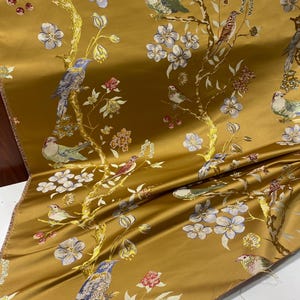 May include: Mustard-colored fabric with a detailed pattern of birds, flowers, and branches. The design features birds in shades of blue, green, and brown perched among white and blue flowers and red berries. The fabric has a satin sheen.