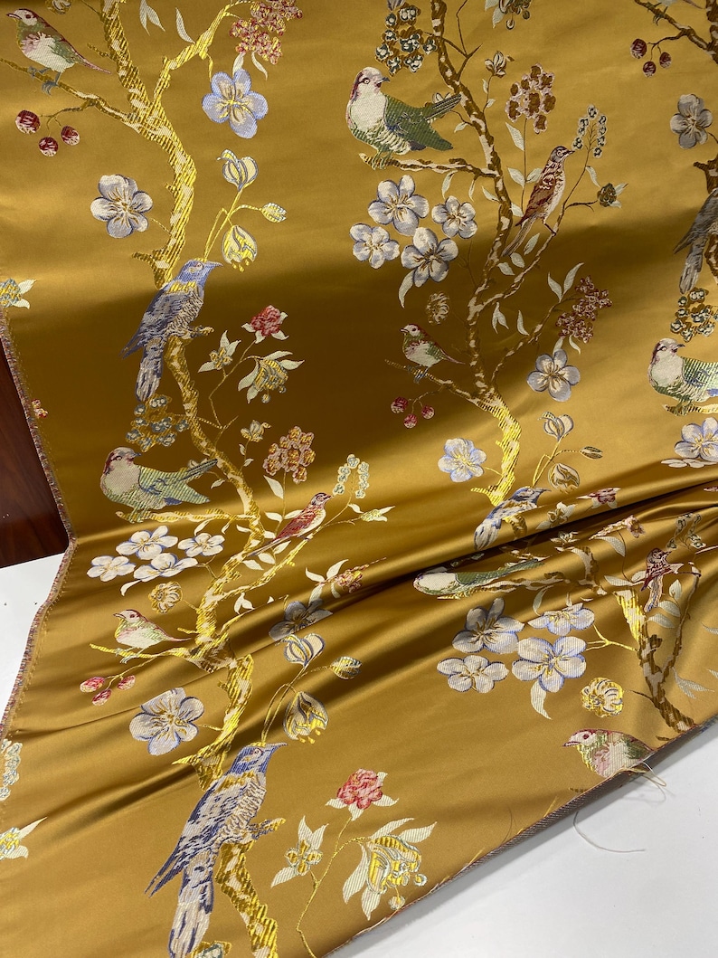 May include: Mustard-colored fabric with a detailed pattern of birds, flowers, and branches. The design features birds in shades of blue, green, and brown perched among white and blue flowers and red berries. The fabric has a satin sheen.