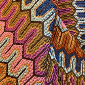 May include: Close-up of a colorful textile with a repeating geometric pattern. The design features a honeycomb-like structure with wavy lines in shades of orange, pink, blue, and brown. The fabric has a textured appearance.