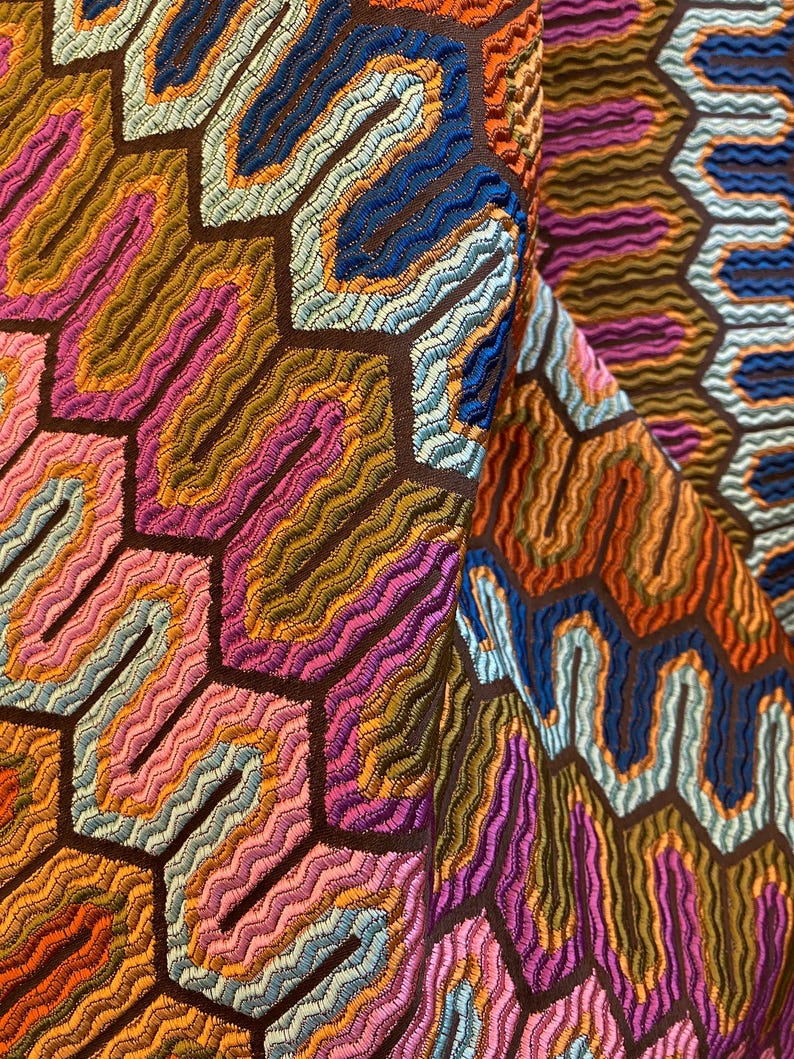 May include: Close-up of a colorful textile with a repeating geometric pattern. The design features a honeycomb-like structure with wavy lines in shades of orange, pink, blue, and brown. The fabric has a textured appearance.