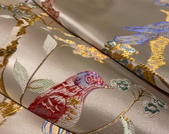May include: Close-up of a folded fabric with a detailed bird and floral pattern. The fabric has a light beige background with embroidered birds in red, blue, and gold, and floral elements in gold and green. The fabric has a satin sheen.