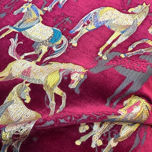 Puede incluir: Una tela de terciopelo rojo intenso con un patrón tejido repetitivo de caballos en tonos dorados, azules y grises. Los caballos aparecen en varias poses dinámicas, con intrincados detalles geométricos sobre el fondo rojo texturizado.