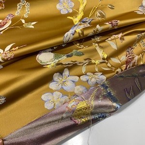 May include: Mustard yellow fabric with an embroidered floral and bird design. The fabric features detailed embroidery of birds, flowers, and branches in shades of blue, white, and brown. The fabric has a satin sheen and a contrasting border.