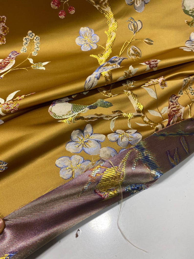 May include: Mustard yellow fabric with an embroidered floral and bird design. The fabric features detailed embroidery of birds, flowers, and branches in shades of blue, white, and brown. The fabric has a satin sheen and a contrasting border.