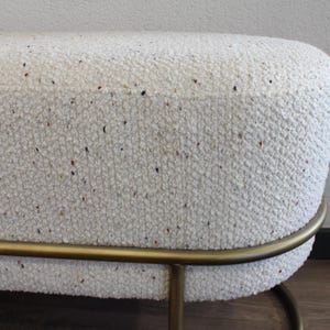 Puede incluir: Banco ovalado blanco con un marco de metal dorado. El banco tiene un tejido texturizado con pequeñas motas blancas, rojas y negras.