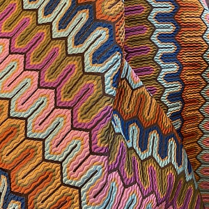 May include: Close-up of a colorful fabric with a geometric pattern. The design features a repeating honeycomb-like structure with wavy lines in shades of orange, blue, pink, and brown. The fabric has a textured appearance.