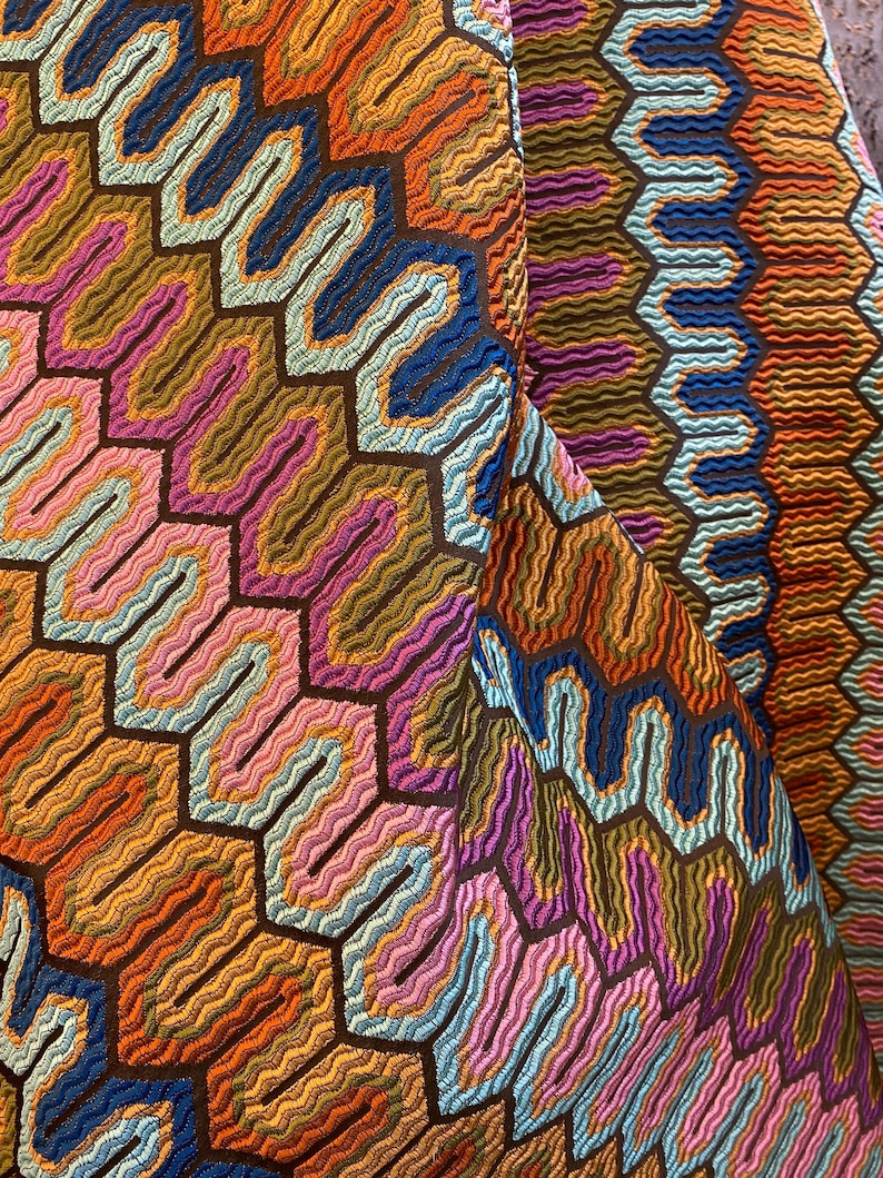 May include: Close-up of a colorful fabric with a geometric pattern. The design features a repeating honeycomb-like structure with wavy lines in shades of orange, blue, pink, and brown. The fabric has a textured appearance.