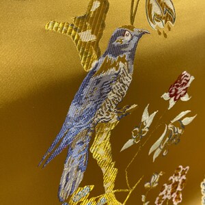May include: A golden yellow fabric featuring an intricate woven pattern of a blue bird perched on a branch. The design includes light blue flowers, white floral buds, and small red blossoms, all rendered in a detailed, textured jacquard weave.