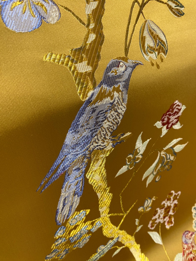 May include: A golden yellow fabric featuring an intricate woven pattern of a blue bird perched on a branch. The design includes light blue flowers, white floral buds, and small red blossoms, all rendered in a detailed, textured jacquard weave.