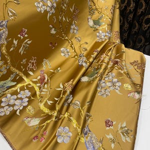May include: A golden yellow fabric featuring a woven pattern of birds perched on flowering branches. The design includes light blue, white, and red floral blossoms along with birds in shades of blue, green, and brown, all set against a smooth, lustrous background.