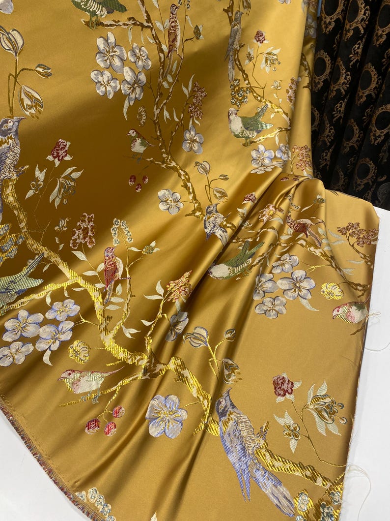 May include: A golden yellow fabric featuring a woven pattern of birds perched on flowering branches. The design includes light blue, white, and red floral blossoms along with birds in shades of blue, green, and brown, all set against a smooth, lustrous background.