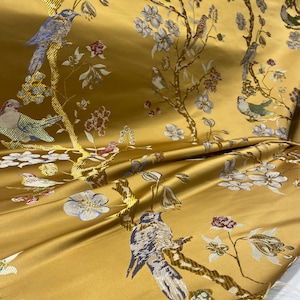 May include: Mustard yellow fabric with a detailed floral and bird pattern. The design features birds perched on branches with white and blue flowers and red accents. The fabric has a satin sheen and is folded, showing the texture and pattern.