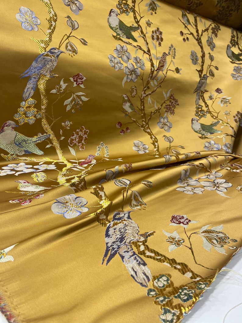 May include: Mustard yellow fabric with a detailed floral and bird pattern. The design features birds perched on branches with white and blue flowers and red accents. The fabric has a satin sheen and is folded, showing the texture and pattern.