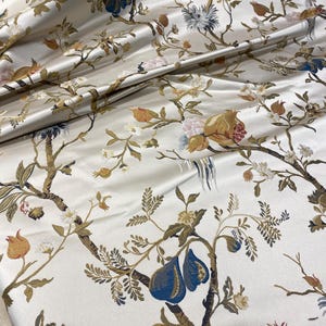 May include: Ivory-colored fabric with a detailed floral pattern. The design features branches with leaves, flowers, and fruits in shades of blue, brown, yellow, and pink. The fabric has a satin sheen.