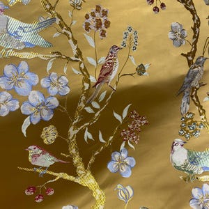 May include: Close-up of a golden fabric with an embroidered design featuring birds, flowers, and branches. The birds are in shades of brown, blue, and white, while the flowers are blue and the branches are gold.