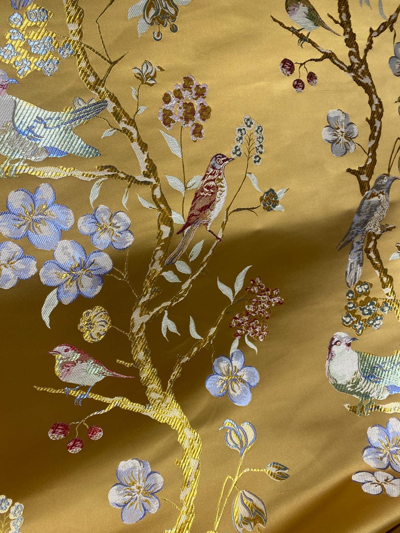 May include: Close-up of a golden fabric with an embroidered design featuring birds, flowers, and branches. The birds are in shades of brown, blue, and white, while the flowers are blue and the branches are gold.