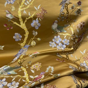 May include: Mustard-colored fabric with an embroidered design of birds perched on golden branches with blue and white flowers. The fabric has a satin sheen and a detailed, repeating pattern. The birds are in shades of red, green, and gray.
