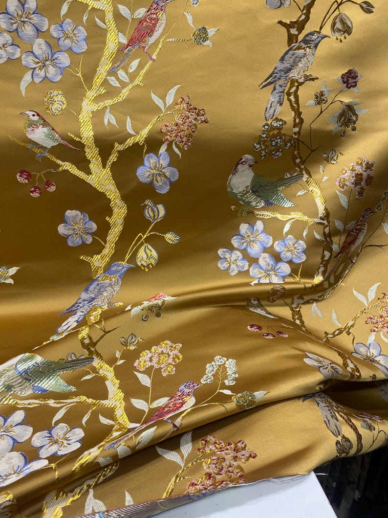 May include: Mustard-colored fabric with an embroidered design of birds perched on golden branches with blue and white flowers. The fabric has a satin sheen and a detailed, repeating pattern. The birds are in shades of red, green, and gray.