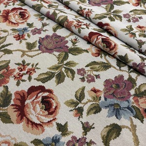 Puede incluir: Primer plano de tela floral con fondo crema. La tela presenta un patrón repetido de rosas en tonos rojos, rosas y morados, con hojas y tallos verdes. La tela está doblada, mostrando la textura y el patrón.