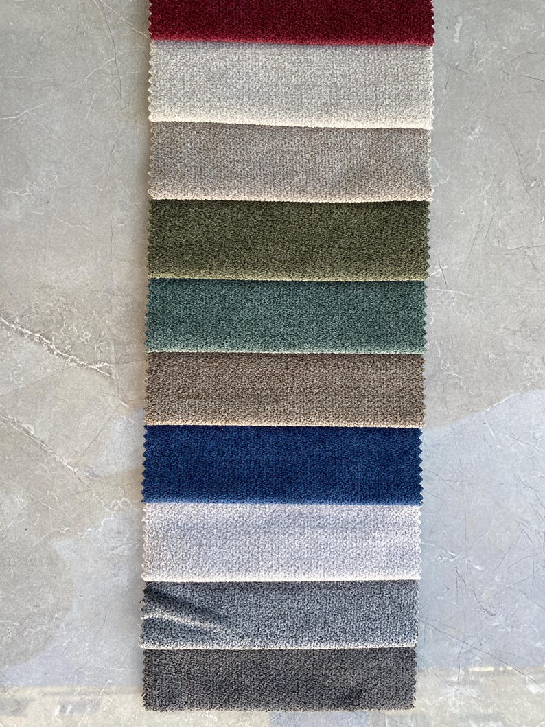 Op de afbeelding: Een verticale weergave van stofstalen in diverse kleuren, waaronder bordeaux, cr&egrave;me, grijs, olijfgroen, blauwgroen, bruin, marineblauw, lichtgrijs en antraciet. De stalen hebben een getextureerd uiterlijk, waarschijnlijk voor bekleding of interieurdecoratie.