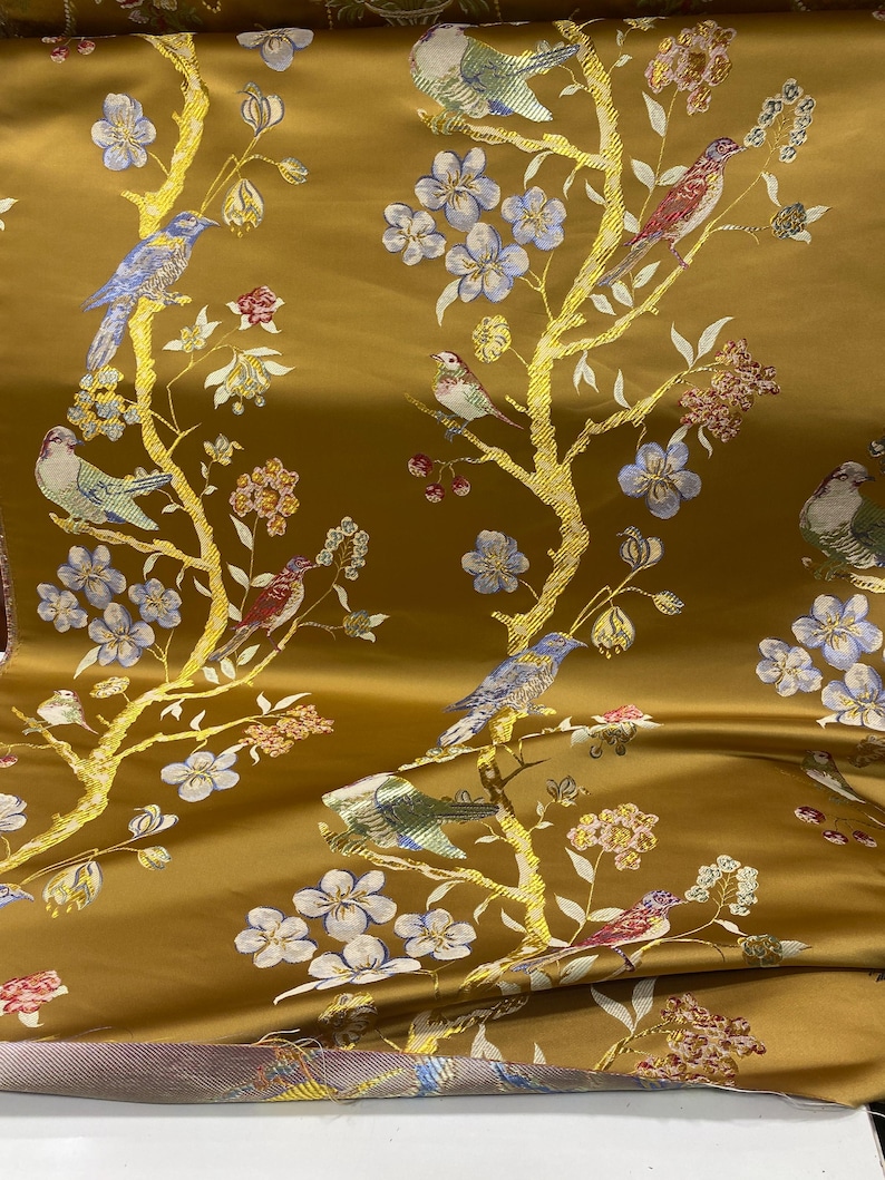 May include: Gold-colored fabric with a repeating pattern of birds perched on flowering branches. The birds are in shades of blue, red, and green, with the branches in gold. The flowers are in shades of blue, pink, and white.