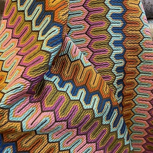 May include: Close-up of a colorful fabric with a geometric pattern. The design features a repeating honeycomb-like structure with wavy lines in shades of orange, blue, pink, and green. The fabric has a textured appearance.