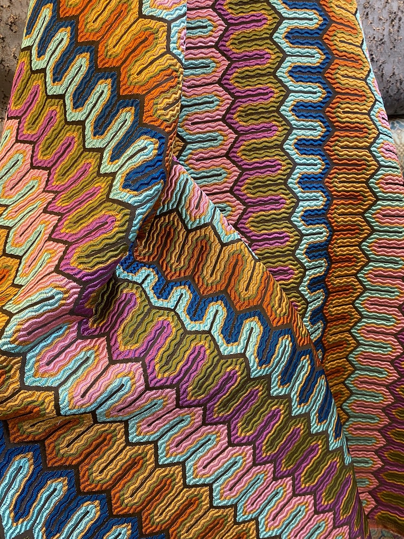 May include: Close-up of a colorful fabric with a geometric pattern. The design features a repeating honeycomb-like structure with wavy lines in shades of orange, blue, pink, and green. The fabric has a textured appearance.