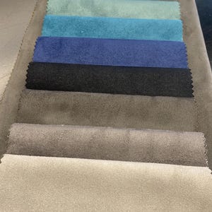 May include: A selection of fabric swatches in various colors, including beige, gray, teal, blue, and black. The samples are arranged in a row, showcasing the different textures and shades available for upholstery or design projects.