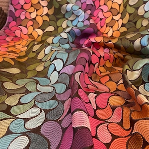 Op de afbeelding: Close-up van een stof met een herhalend patroon van kleurrijke, druppelvormige ontwerpen. De stof heeft een donkerbruine achtergrond met de druppels in tinten roze, oranje, geel, groen, blauw en paars. De stof is getextureerd en heeft een lichte glans.