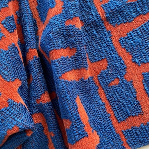 Könnte beinhalten: Nahaufnahme eines strukturierten Stoffes mit einem sich wiederholenden Muster. Der Stoff weist eine lebendige Kombination aus Blau und Orange auf. Das Design besteht aus orangefarbenen geometrischen Formen auf blauem Hintergrund, was einen auffälligen Kontrast erzeugt.