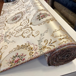 Könnte beinhalten: Eine Stoffrolle in Cremefarben mit einem sich wiederholenden Muster aus goldenen Schnörkeln, Blumensträußen in Rosa und Grün sowie ovalen Rahmen. Das Textil hat eine glatte, satinartige Oberfläche mit einer ausgefransten Kante und Webstruktur.