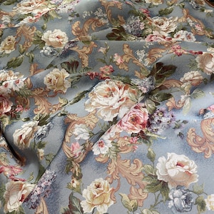 Könnte beinhalten: Ein Stoff mit hellblauem Hintergrund und einem sich wiederholenden Muster aus cremefarbenen, rosafarbenen und weißen Rosen, grünen Blättern und beigen Schnörkeln. Das Textil hat ein strukturiertes Gewebe und ist in Falten gelegt.