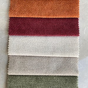 Op de afbeelding: Een verticale weergave van stoffen stalen in verschillende kleuren. De stalen omvatten tinten goud, oranje, bordeauxrood, gebroken wit, grijs, olijfgroen en blauwgroen. De stof heeft een getextureerde, geluste uitstraling.