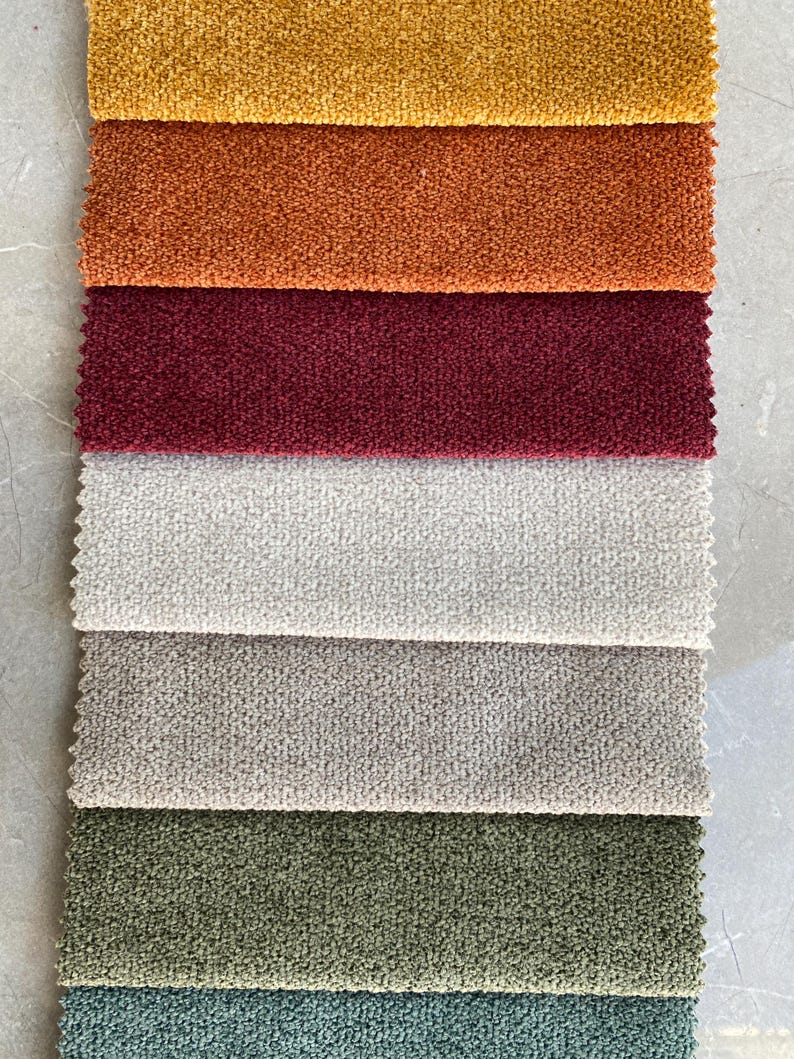 Op de afbeelding: Een verticale weergave van stoffen stalen in verschillende kleuren. De stalen omvatten tinten goud, oranje, bordeauxrood, gebroken wit, grijs, olijfgroen en blauwgroen. De stof heeft een getextureerde, geluste uitstraling.