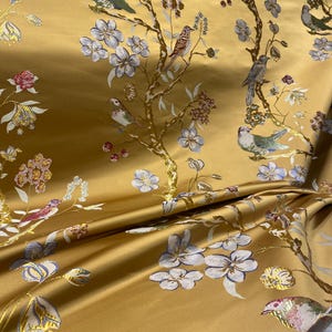 May include: A golden yellow fabric featuring a woven pattern of birds perched on flowering branches. The design includes light blue, pink, and white blossoms, along with various birds in shades of gray, green, and blue, all set against a smooth, lustrous background.