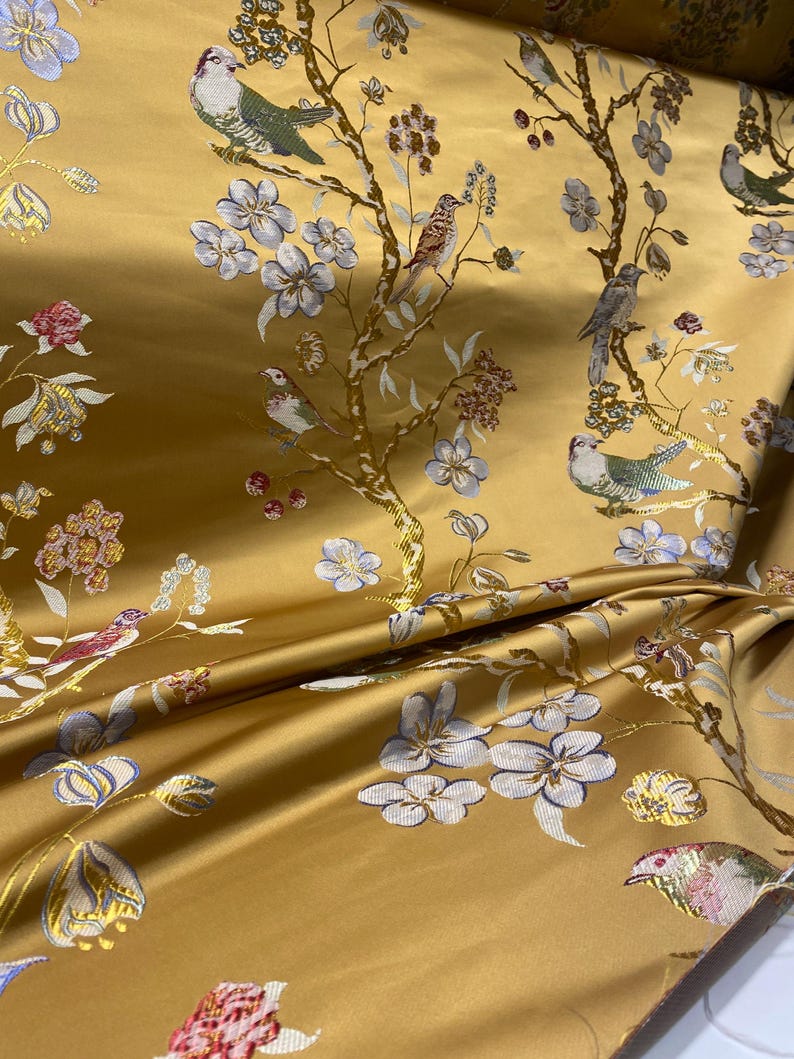 May include: A golden yellow fabric featuring a woven pattern of birds perched on flowering branches. The design includes light blue, pink, and white blossoms, along with various birds in shades of gray, green, and blue, all set against a smooth, lustrous background.