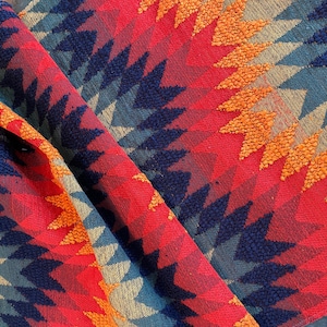 May include: Close-up of a folded textile with a geometric pattern. The design features a repeating chevron pattern in shades of red, orange, blue, and tan. The fabric has a textured appearance.