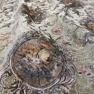 Könnte beinhalten: Nahaufnahme eines salbeigrünen Stoffes mit einem verzierten Blumendesign. Der Stoff weist detaillierte Muster aus Blumen, Wirbeln und dekorativen Rahmen in Gold-, Braun- und Rosatönen auf. Die Gesamtstruktur wirkt weich und luxuriös.