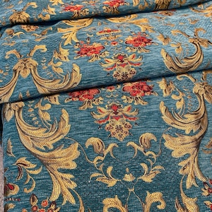 May include: Close-up of a teal fabric with a detailed gold and red floral pattern. The fabric has a textured appearance with a repeating design of stylized flowers and ornate scrollwork, creating a luxurious look.