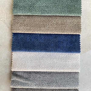 Op de afbeelding: Een verticale weergave van stofstalen in verschillende kleuren. De stalen omvatten tinten groen, blauw, bruin en grijs. De stoffen hebben een getextureerde, geluste uitstraling, wat wijst op een pluche of boucl&eacute; materiaal, geschikt voor bekleding of interieurdecoratie.