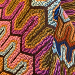May include: Close-up of a colorful fabric with a geometric pattern. The design features a series of interconnected hexagons filled with wavy, embroidered lines in shades of pink, orange, green, and blue, set against a dark brown background.