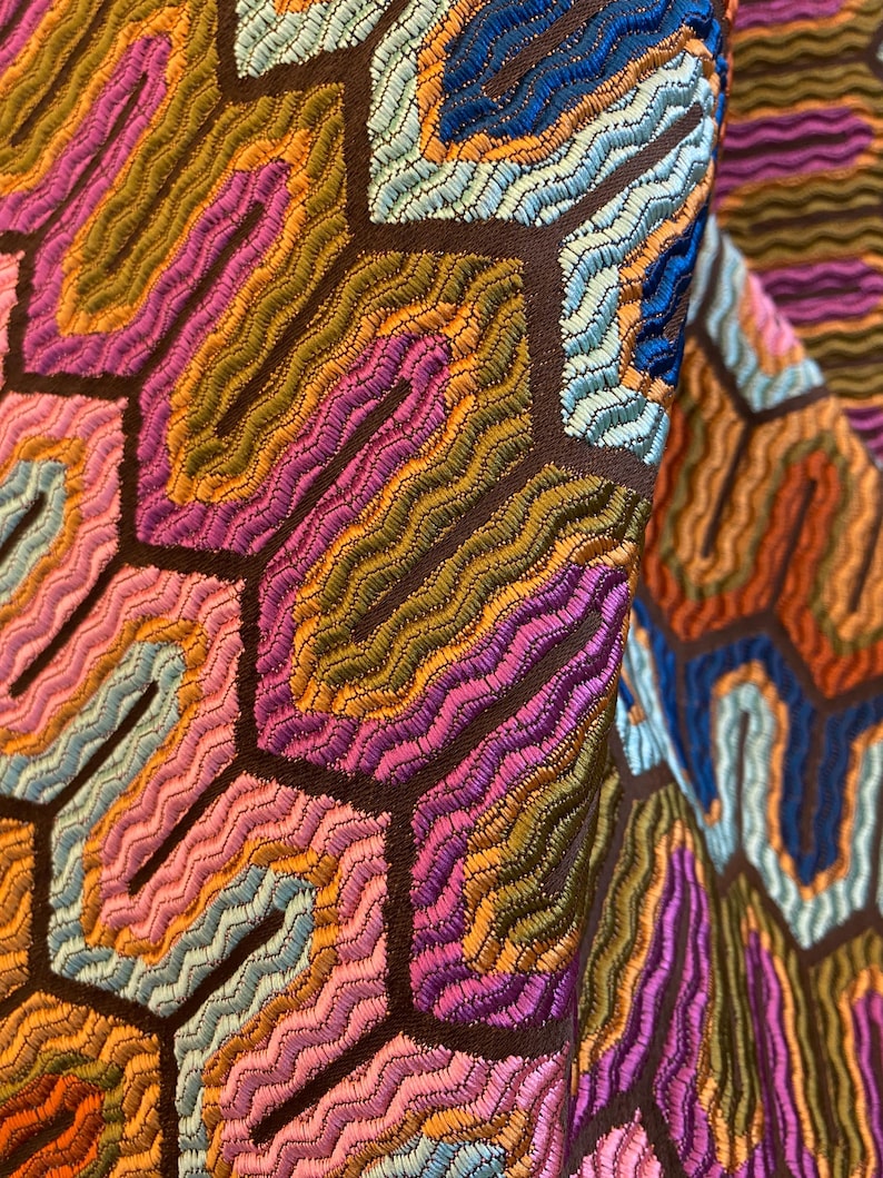 May include: Close-up of a colorful fabric with a geometric pattern. The design features a series of interconnected hexagons filled with wavy, embroidered lines in shades of pink, orange, green, and blue, set against a dark brown background.