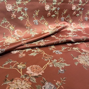 May include: A close-up of a fabric with a floral pattern. The fabric is a reddish-brown color with embroidered flowers in shades of pink, blue, and green. The flowers are arranged in a repeating pattern across the fabric.