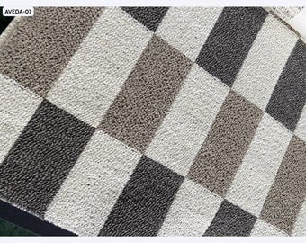 Puede incluir: Una alfombra con diseño geométrico con un diseño de tablero de ajedrez. El patrón de la alfombra consiste en cuadrados y rectángulos alternos en tonos de gris, blanco y marrón. La alfombra tiene una superficie texturizada.