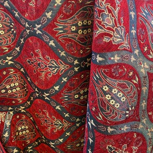 May include: Close-up of a red fabric with an intricate pattern. The design features floral and geometric shapes in gold, black, and dark red. The fabric has a textured appearance, suggesting a woven material, possibly for upholstery or drapery.