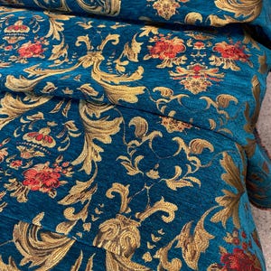 Könnte beinhalten: Nahaufnahme eines gefalteten, türkisfarbenen Stoffes mit einem verzierten goldenen und roten Blumenmuster. Das Design zeigt wirbelnde goldene Blätter und rote Blumengruppen, die ein luxuriöses und strukturiertes Aussehen erzeugen. Der Stoff wird wahrscheinlich für Polster oder Dekorationszwecke verwendet.